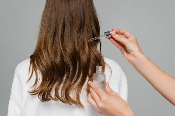 hair stylist applying serum on hair of young woman isolated on grey