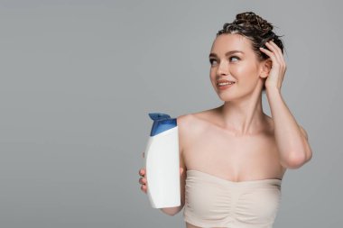 happy young woman with wet foamy hair holding bottle with shampoo isolated on grey  clipart