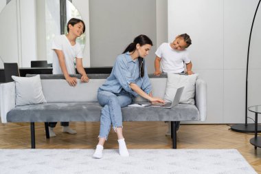 Kulaklıklı anne evde Asyalı çocukların yanında dizüstü bilgisayar kullanıyor. 