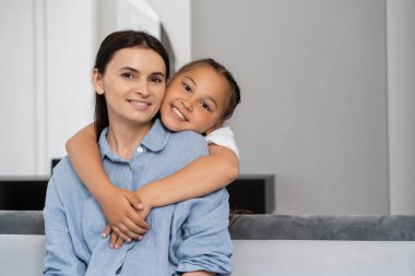 Neşeli Asyalı kız annesine sarılıyor ve evde kameraya bakıyor. 