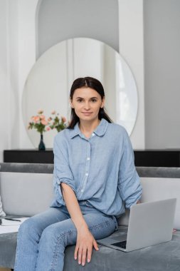 Esmer serbest çalışan oturma odasındaki koltukta dizüstü bilgisayarın yanındaki kameraya bakıyor. 