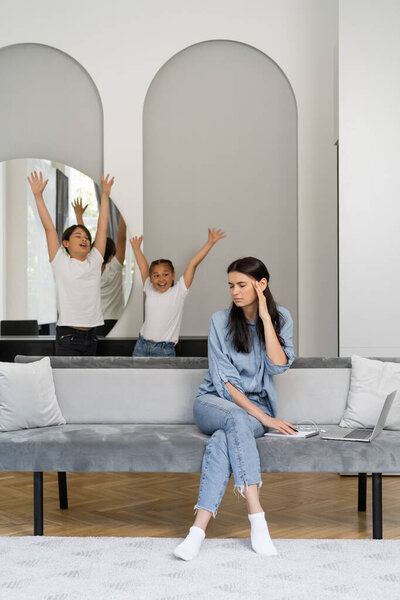 Tired woman sitting near laptop and cheerful asian kids at home 