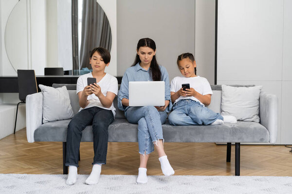 Woman using laptop near asian children with smartphones at home 
