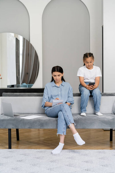 Freelancer writing on notebook near laptop and asian daughter with smartphone at home 