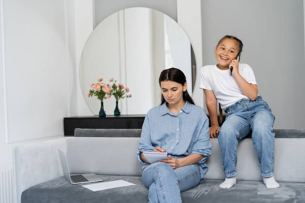 Positive asian kid talking on smartphone near mother with notebook and laptop at home 