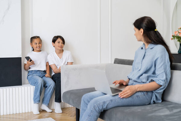 Woman with laptop looking at cheerful asian kids with mobile phone at home 