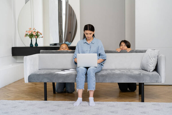 Woman working on laptop near sly asian kids behind couch at home 