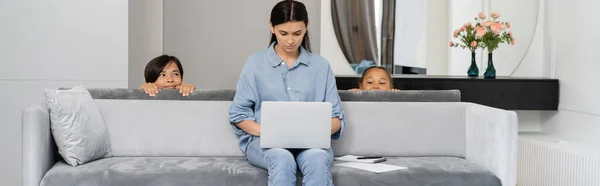 Sly asian kids looking at mom using laptop on couch at home, banner