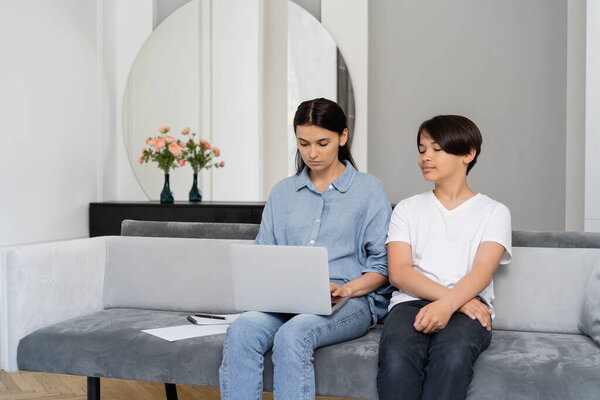 Asian boy sitting near mother using laptop on couch at home 