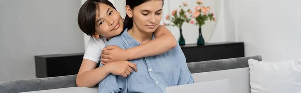 Asian kid hugging mom looking at laptop on couch at home, banner