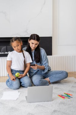 Mother using smartphone and laptop near asian daughter with apple and color pencils at home  clipart