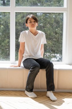 Preteen asian boy sitting on windowsill at home  clipart