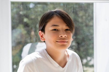 Portrait of preteen asian boy smiling at camera near window at home  clipart