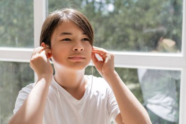 Preteen asian child using earphones near window at home  clipart