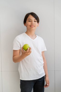 Smiling asian boy holding ripe apple near wall at home  clipart