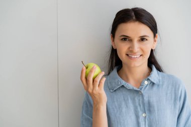 Gülümseyen esmer kadın tişörtlü, duvarın yanında elma tutuyor. 