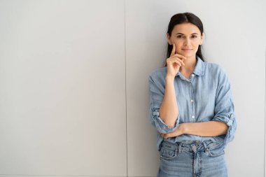 Brunette woman in shirt and jeans looking at camera near grey wall  clipart