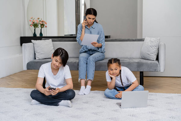 Asian children using devices while mother talking on smartphone and holding paper at home 