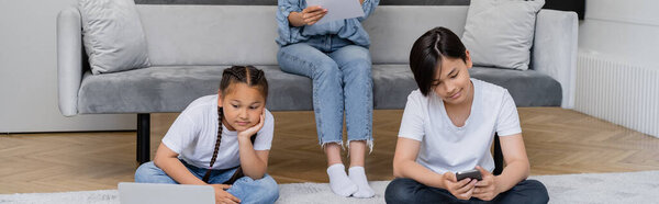 Asian children using gadgets near mom with paper on couch at home, banner 