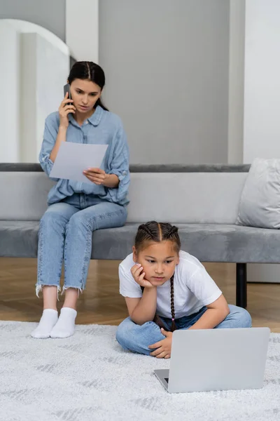 Asian kid watching cartoons on laptop while mother talking on smartphone and holding paper at home