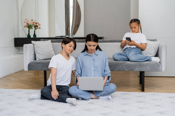 Woman using laptop near asian son and daughter with smartphone at home 