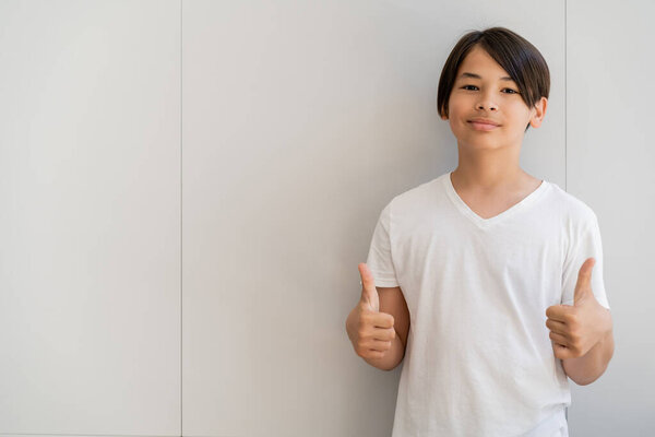 Preteen asian boy showing like gesture near wall at home 