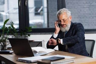 Gözlüklü olgun bir işadamı akıllı telefondan konuşuyor ve ofiste zamanı kontrol ediyor. 