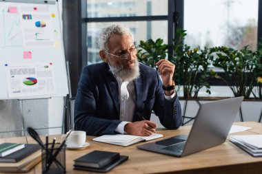 Gözlüklü orta yaşlı işadamı deftere yazı yazıyor ve ofisteki kağıtların yanında dizüstü bilgisayar kullanıyor. 