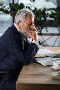 Gözlüklü orta yaşlı işadamı dizüstü bilgisayarın yanında oturuyor ve ofiste not defteri var. 