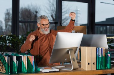 Excited grey haired manager looking at computer monitor near papers and notebooks in office clipart