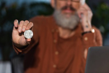 KYIV, UKRAINE - 17 Kasım 2022: bulanık arkaplandaki sakallı iş adamının elindeki gümüş bitcoin 'in seçici odağı