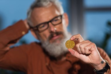 KYIV, UKRAINE - 17 Kasım 2022: Bulanık arkaplanda olgun ve sakallı iş adamının elindeki altın bitcoin 'in seçici odağı