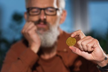 KYIV, UKRAINE - 17 Kasım 2022: Bulanık arkaplandaki düşünceli sakallı iş adamının elinde altın bitcoin 'in seçici odağı