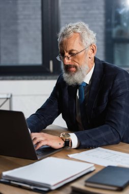 Gözlüklü ve takım elbiseli sakallı bir adam laptopta ofisteki defterlerin yanında daktilo yazıyor.