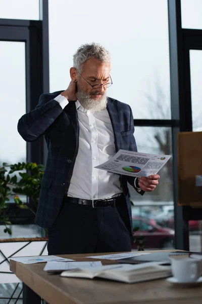 Middle aged manager holding paper with charts near working table in office