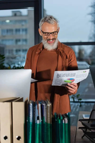正の灰色の髪のビジネスマンは 夕方にオフィスでコンピュータの近くのチャートと文書を見て — ストック写真
