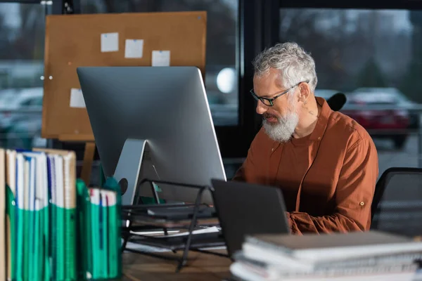 Mature businessman in eyeglasses working near computers and blurred documents in office