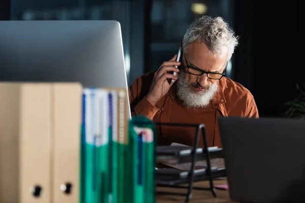 灰色头发的商人戴着眼镜 在电脑前用智能手机交谈 晚上在办公室里的文件夹模糊不清 — 图库照片