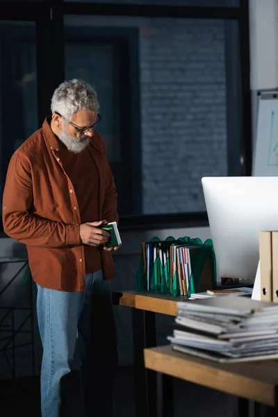 灰色头发的商人站在办公室里的文件和计算机监视器旁边 手里拿着笔记本 — 图库照片