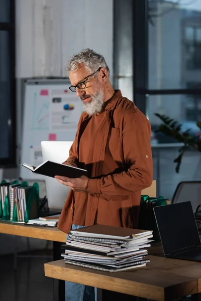 一个戴眼镜的大胡子男子站在堆满文件的工作场所 手里拿着笔记本 — 图库照片