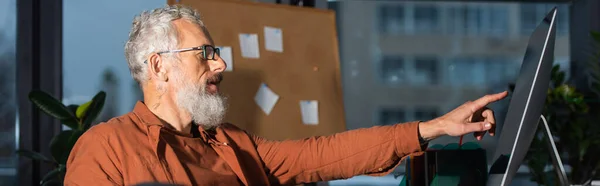 side view of bearded businessman pointing at computer monitor while working in office, banner