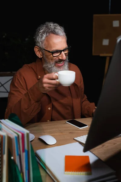 带着咖啡杯的快乐商人坐在智能手机旁的工作场所 电脑显示器模糊不清 — 图库照片