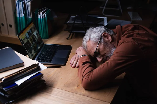 tired grey haired businessman sleeping near laptop and stack of documents in dark office
