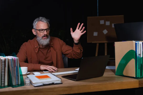 stock image displeased businessman gesturing during video chat on laptop near copybooks in office in evening