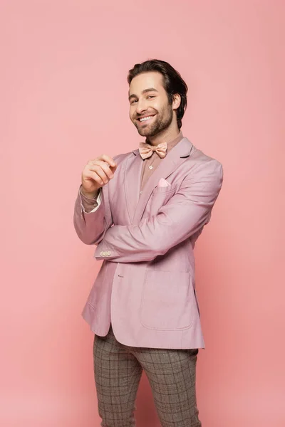 Smiling host of event in jacket and bow tie looking at camera on pink background