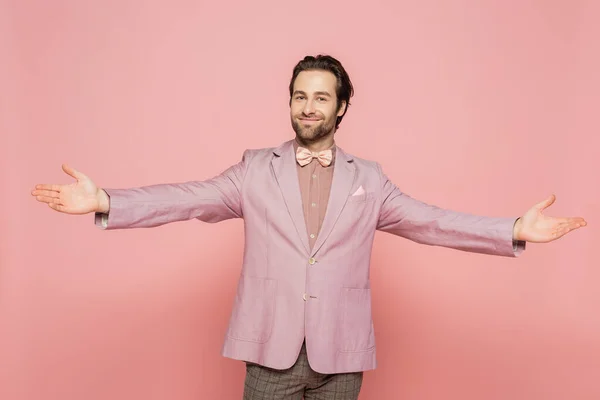 Positive host of event in bow tie and jacket outstretching hands isolated on pink
