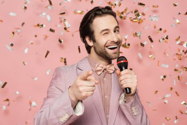 Positive host of event holding microphone and winking at camera under confetti on pink background  clipart
