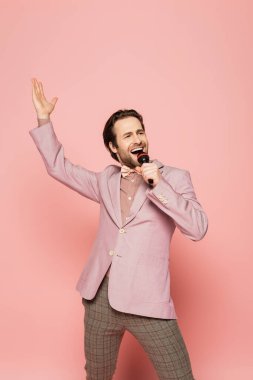 Cheerful host of event singing while holding microphone on pink background  clipart