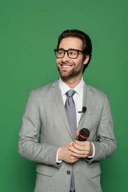 happy news anchor in suit and eyeglasses holding microphone isolated on green  clipart