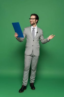full length of cheerful news anchor in suit and eyeglasses holding clipboard while gesturing on green  clipart